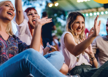 Four adults sitting down at a concert cheering with their hands in the ai