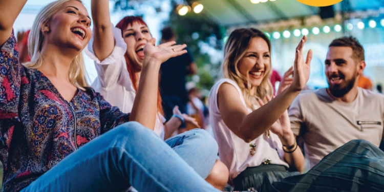 Four adults sitting down at a concert cheering with their hands in the ai